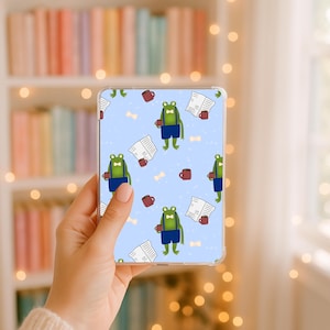 May include: A clear protective case for a tablet or e-reader featuring a repeating pattern on a light blue background. The design includes green frogs wearing blue shorts and white bow ties, holding dark red mugs, alongside open newspapers.