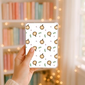May include: A clear rectangular phone case featuring a repeating pattern of brown coffee cups with heart-shaped latte art, small green leafy sprigs, and tiny light purple hearts printed on a white background, held in a hand.