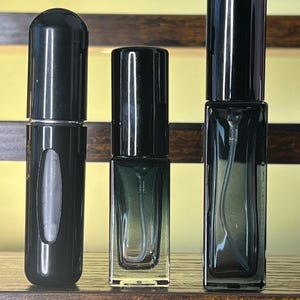 May include: Three empty glass spray bottles with black caps. The left bottle is cylindrical with a small viewing window. The middle and right bottles are rectangular with a dark green gradient tint and visible internal spray tubes.
