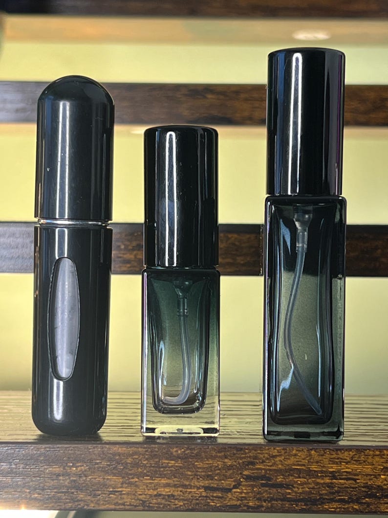 May include: Three empty glass spray bottles with black caps. The left bottle is cylindrical with a small viewing window. The middle and right bottles are rectangular with a dark green gradient tint and visible internal spray tubes.