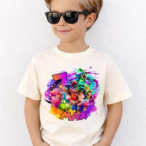 Puede incluir: Una camiseta de manga corta de color crema con un estampado gr&aacute;fico colorido de varios personajes de videojuegos, incluidos Mario, Luigi, Yoshi y Bowser, junto con el n&uacute;mero 7 y el nombre MAX en letras llamativas, vibrantes y multicolores.