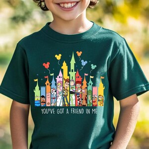 May include: Dark green t-shirt featuring a colorful graphic of cartoon characters and castles. The text "YOU'VE GOT A FRIEND IN ME" is printed at the bottom. The design includes balloons in the sky.