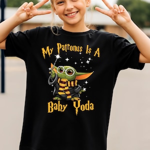 May include: Black t-shirt with the text "My Patronus Is A Baby Yoda" in gold lettering. The graphic features a cartoon Baby Yoda character with glasses, a wand, and a scarf. The shirt is worn by a person.