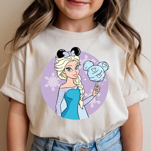 May include: A beige t-shirt featuring a cartoon illustration of a princess with blonde hair, wearing a blue dress and a black bow. She is holding a blue cotton candy lollipop. The background is a purple circle with snowflakes.