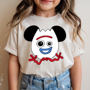 May include: A beige t-shirt featuring a cartoon character design. The character has a white face with black ears, two black eyes, a red smile, and a blue mouth. Red squiggly lines form the character's arms and legs. The shirt is worn by a person.