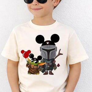 May include: Cream-colored t-shirt featuring a cartoon illustration of Mickey Mouse as The Mandalorian and Grogu. Mickey Mouse is wearing a Mandalorian helmet and Grogu is holding red balloons. The shirt is for children.