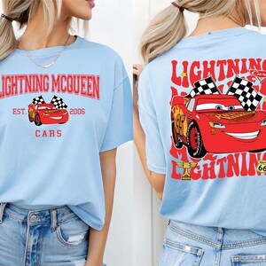 May include: Light blue t-shirt featuring Lightning McQueen graphics. The front design includes the text "Lightning McQueen Cars Est. 2006" with a red car and checkered flags. The back design showcases a larger image of the red car with checkered flags and the text "Lightning Cars".