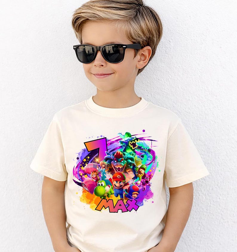 Puede incluir: Una camiseta de manga corta de color crema con un estampado gr&aacute;fico colorido de varios personajes de videojuegos, incluidos Mario, Luigi, Yoshi y Bowser, junto con el n&uacute;mero 7 y el nombre MAX en letras llamativas, vibrantes y multicolores.