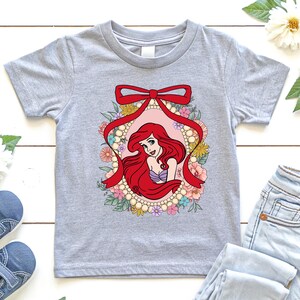 May include: A light gray t-shirt featuring a colorful graphic of a mermaid with red hair, a purple seashell top, and a floral frame. The design includes a red bow and pearl accents. The shirt is a children's size.