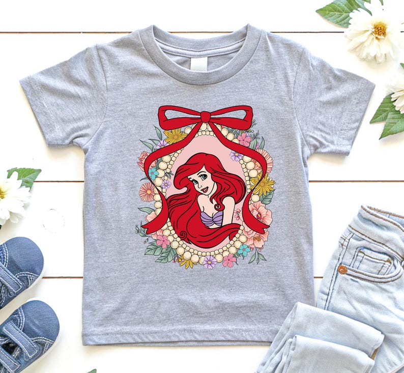 May include: A light gray t-shirt featuring a colorful graphic of a mermaid with red hair, a purple seashell top, and a floral frame. The design includes a red bow and pearl accents. The shirt is a children's size.