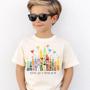 May include: Cream-colored t-shirt featuring a colorful castle design with cartoon characters and balloons. The text "YOU'VE GOT A FRIEND IN ME" is printed below the design. The shirt is worn by a child wearing black sunglasses.