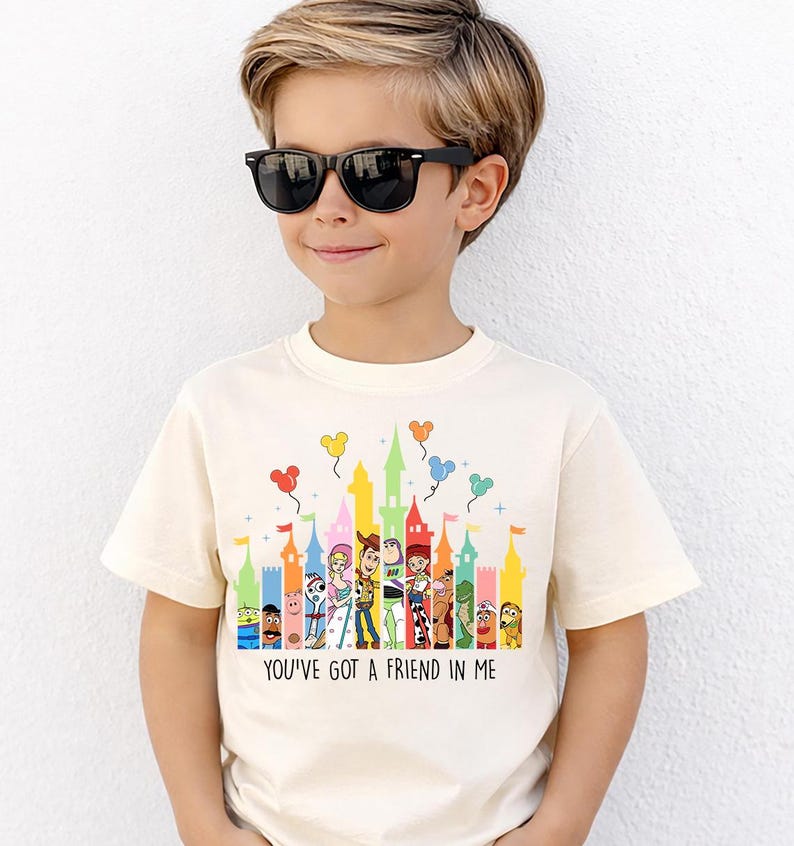 May include: Cream-colored t-shirt featuring a colorful castle design with cartoon characters and balloons. The text "YOU'VE GOT A FRIEND IN ME" is printed below the design. The shirt is worn by a child wearing black sunglasses.