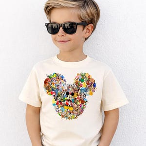 Puede incluir: Una camiseta color crema con un dise&ntilde;o de la cabeza de Mickey Mouse compuesto por varios personajes de Disney. La camiseta es de manga corta y la lleva un ni&ntilde;o con gafas de sol negras.