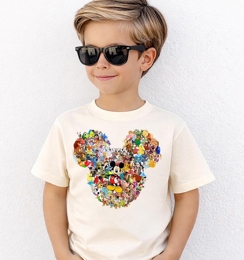 Puede incluir: Una camiseta color crema con un dise&ntilde;o de la cabeza de Mickey Mouse compuesto por varios personajes de Disney. La camiseta es de manga corta y la lleva un ni&ntilde;o con gafas de sol negras.
