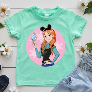 May include: Mint green t-shirt featuring a cartoon illustration of a character with red braids, wearing a black top and holding an ice cream cone. The character is set against a pink circle with snowflake accents, and wears black mouse ears.
