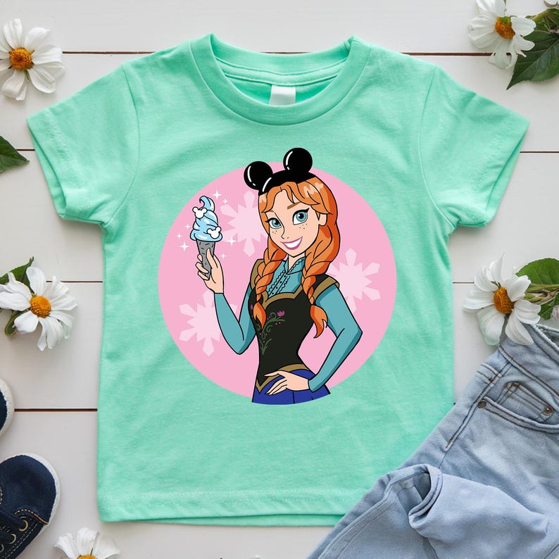 May include: Mint green t-shirt featuring a cartoon illustration of a character with red braids, wearing a black top and holding an ice cream cone. The character is set against a pink circle with snowflake accents, and wears black mouse ears.