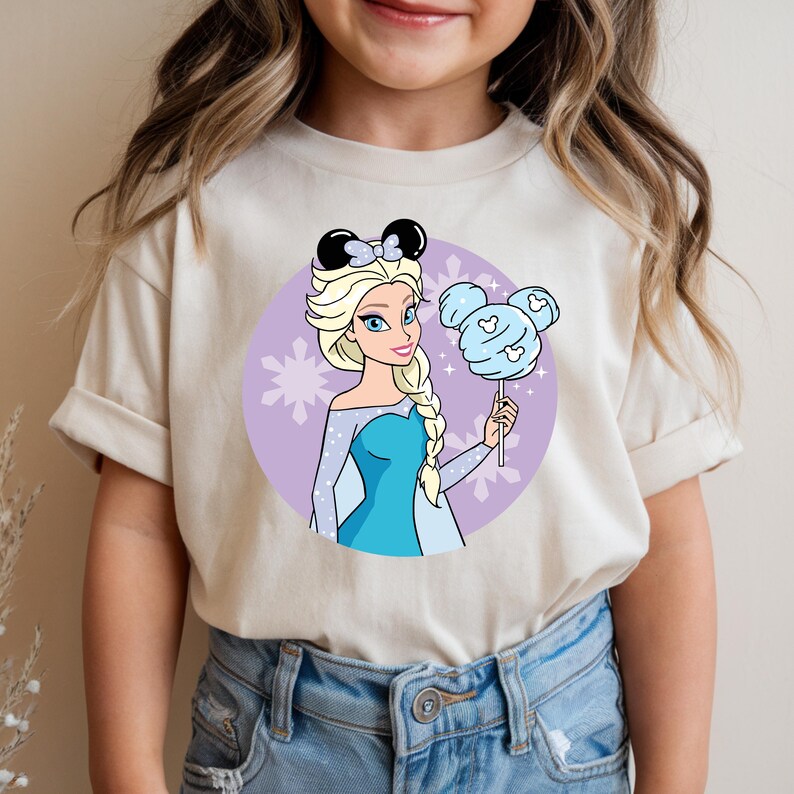 May include: A beige t-shirt featuring a cartoon illustration of a princess with blonde hair, wearing a blue dress and a black bow. She is holding a blue cotton candy lollipop. The background is a purple circle with snowflakes.