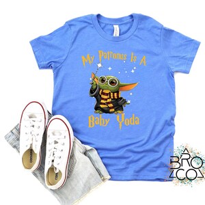 May include: A light blue t-shirt with the text "My Patronus Is A Baby Yoda" in gold. The graphic features a cartoon Baby Yoda character wearing glasses and a scarf. White sneakers and denim shorts are also in the image.