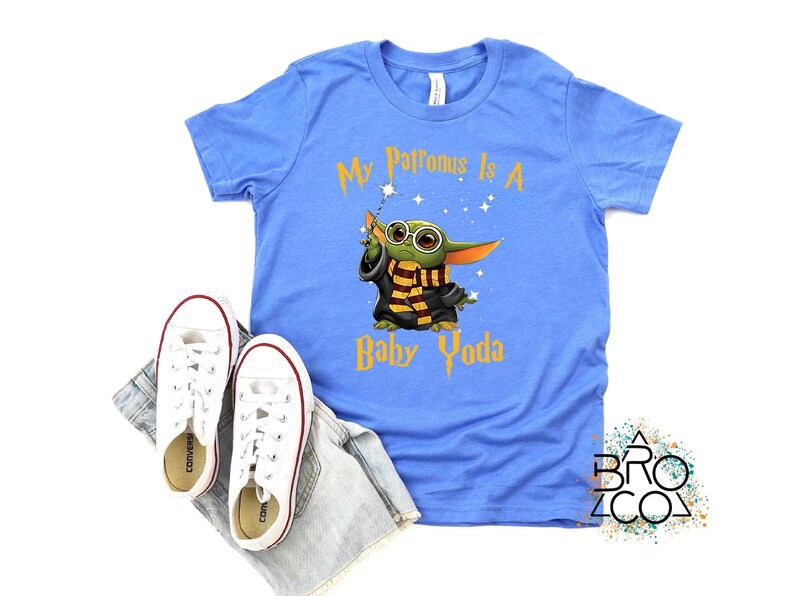 May include: A light blue t-shirt with the text "My Patronus Is A Baby Yoda" in gold. The graphic features a cartoon Baby Yoda character wearing glasses and a scarf. White sneakers and denim shorts are also in the image.
