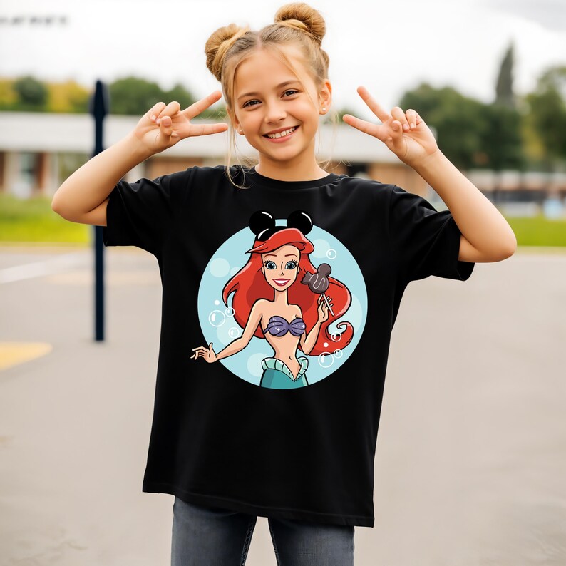 May include: Black t-shirt featuring a cartoon illustration of Ariel from The Little Mermaid, with a Mickey Mouse ear headband. Ariel has red hair, a purple seashell bra, and a teal tail. She is holding a lollipop. The design is set against a blue background.