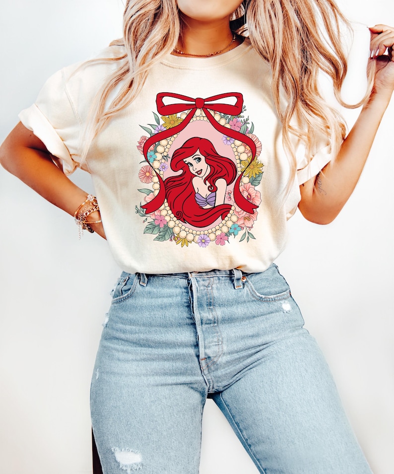 May include: Cream-colored t-shirt featuring a graphic of a red-haired mermaid within a floral and pearl-adorned oval, accented by a red bow. The mermaid is wearing a purple top. The shirt is paired with light blue jeans.