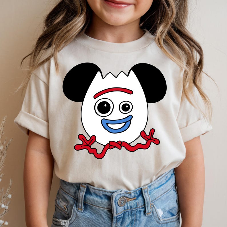 May include: A beige t-shirt featuring a cartoon character design. The character has a white face with black ears, two black eyes, a red smile, and a blue mouth. Red squiggly lines form the character's arms and legs. The shirt is worn by a person.