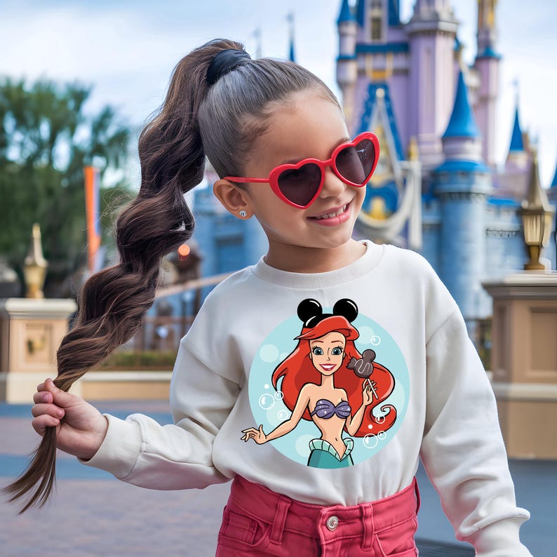 May include: A white sweatshirt featuring a cartoon image of Ariel from The Little Mermaid, with Mickey Mouse ears. The design is set against a light blue background with bubbles. The girl is wearing red heart-shaped sunglasses and pink pants.