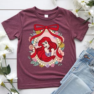 May include: Burgundy t-shirt featuring a cartoon illustration of a red-haired mermaid within a floral and pearl-bordered oval. A red ribbon bow tops the design. The mermaid wears a purple seashell top. The shirt is paired with blue jeans.
