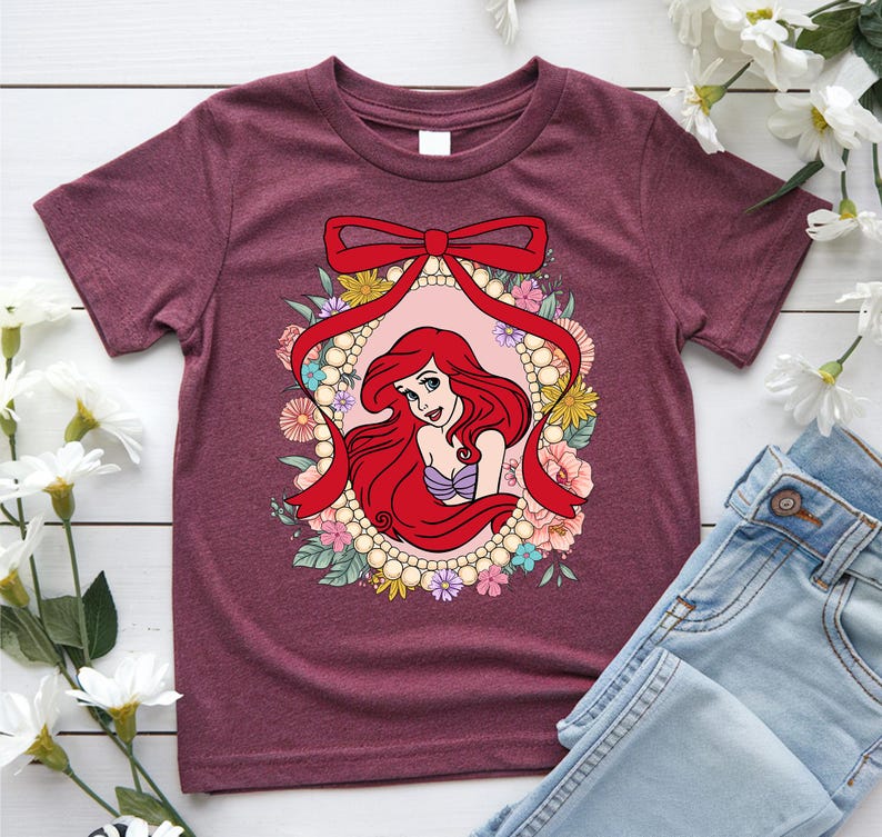 May include: Burgundy t-shirt featuring a cartoon illustration of a red-haired mermaid within a floral and pearl-bordered oval. A red ribbon bow tops the design. The mermaid wears a purple seashell top. The shirt is paired with blue jeans.