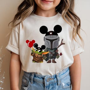 May include: A beige t-shirt featuring a cartoon illustration of The Mandalorian and Grogu, both with Mickey Mouse ears. Grogu holds a red balloon. The Mandalorian is holding a stick. The design is in black, green, brown, and red.