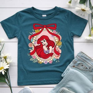 May include: Teal-colored t-shirt featuring a graphic of a red-haired cartoon character within a floral and pearl-adorned frame, accented by a red bow. The character has a purple seashell top. The shirt is paired with light blue jeans.