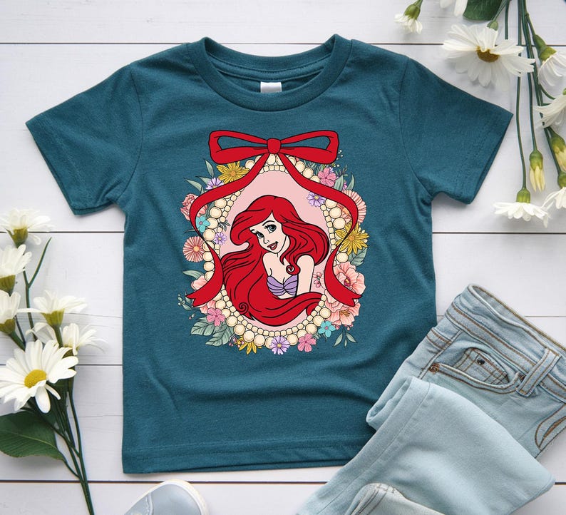 May include: Teal-colored t-shirt featuring a graphic of a red-haired cartoon character within a floral and pearl-adorned frame, accented by a red bow. The character has a purple seashell top. The shirt is paired with light blue jeans.