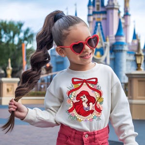 May include: A cream-colored sweatshirt featuring a graphic of Ariel from The Little Mermaid, framed by a red ribbon and floral accents. The design is centered on the front of the long-sleeved top. The girl is wearing red heart-shaped sunglasses and red pants.