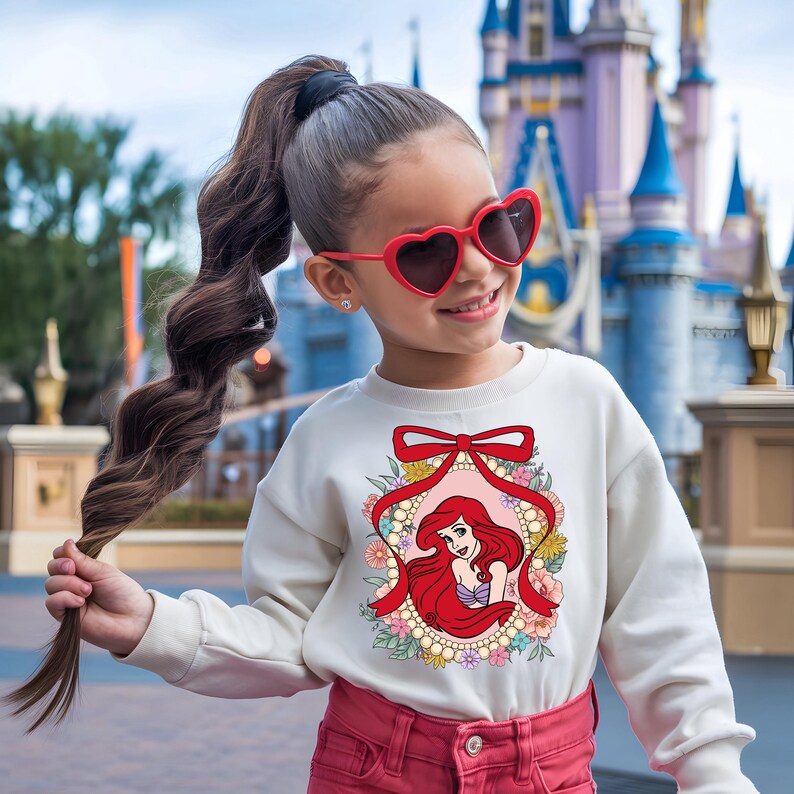 May include: A cream-colored sweatshirt featuring a graphic of Ariel from The Little Mermaid, framed by a red ribbon and floral accents. The design is centered on the front of the long-sleeved top. The girl is wearing red heart-shaped sunglasses and red pants.