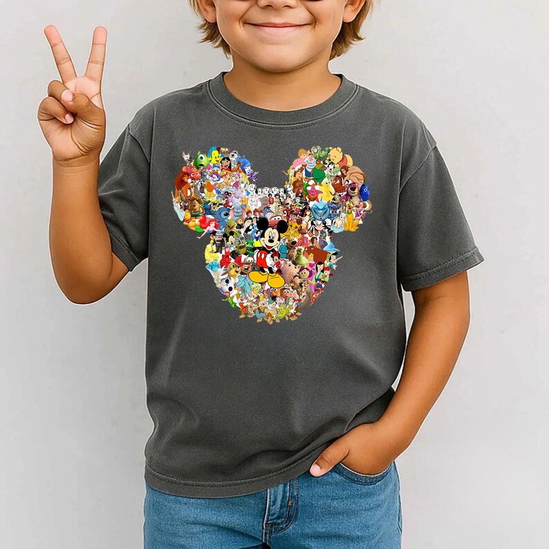 Puede incluir: Camiseta gris oscuro con un dise&ntilde;o de la cabeza de Mickey Mouse compuesto por coloridos personajes de Disney. La camiseta la lleva un ni&ntilde;o que hace el signo de la paz. Es una prenda informal de manga corta.