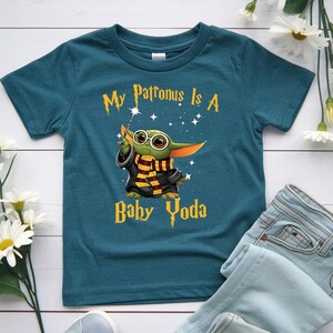 May include: Teal blue t-shirt featuring a graphic of Baby Yoda dressed as a wizard with the text "My Patronus Is A Baby Yoda". The design includes stars and a wand. The shirt is paired with blue jeans.