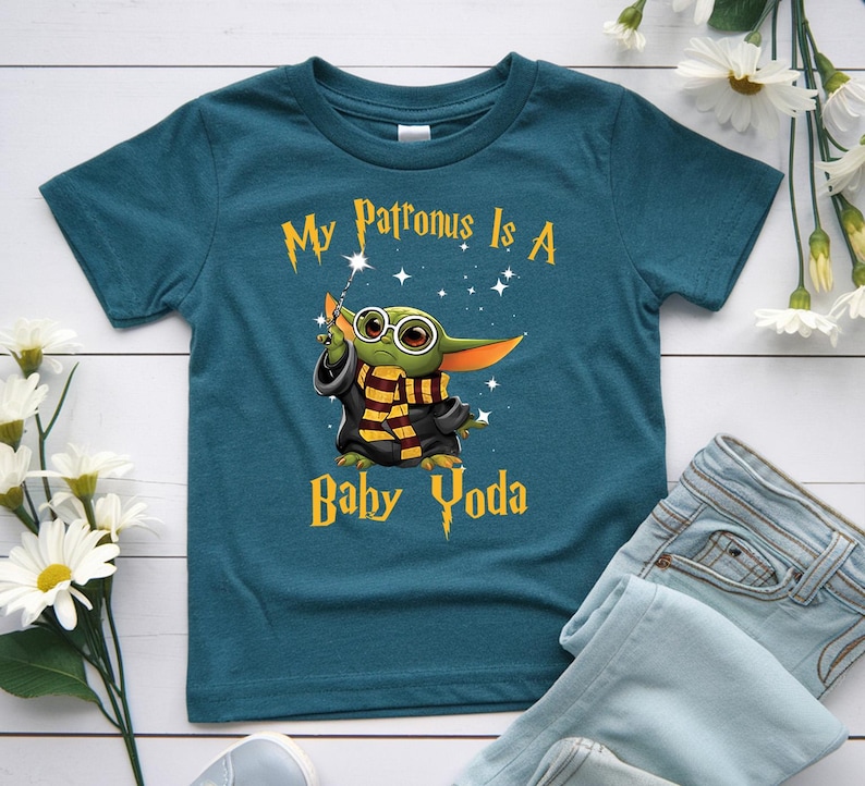 May include: Teal blue t-shirt featuring a graphic of Baby Yoda dressed as a wizard with the text "My Patronus Is A Baby Yoda". The design includes stars and a wand. The shirt is paired with blue jeans.