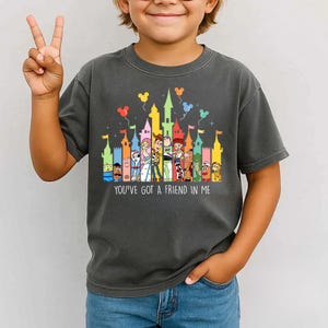 May include: Dark gray t-shirt featuring a colorful castle design with cartoon characters and the text "YOU'VE GOT A FRIEND IN ME". The design includes balloons and flags, creating a playful and whimsical look.