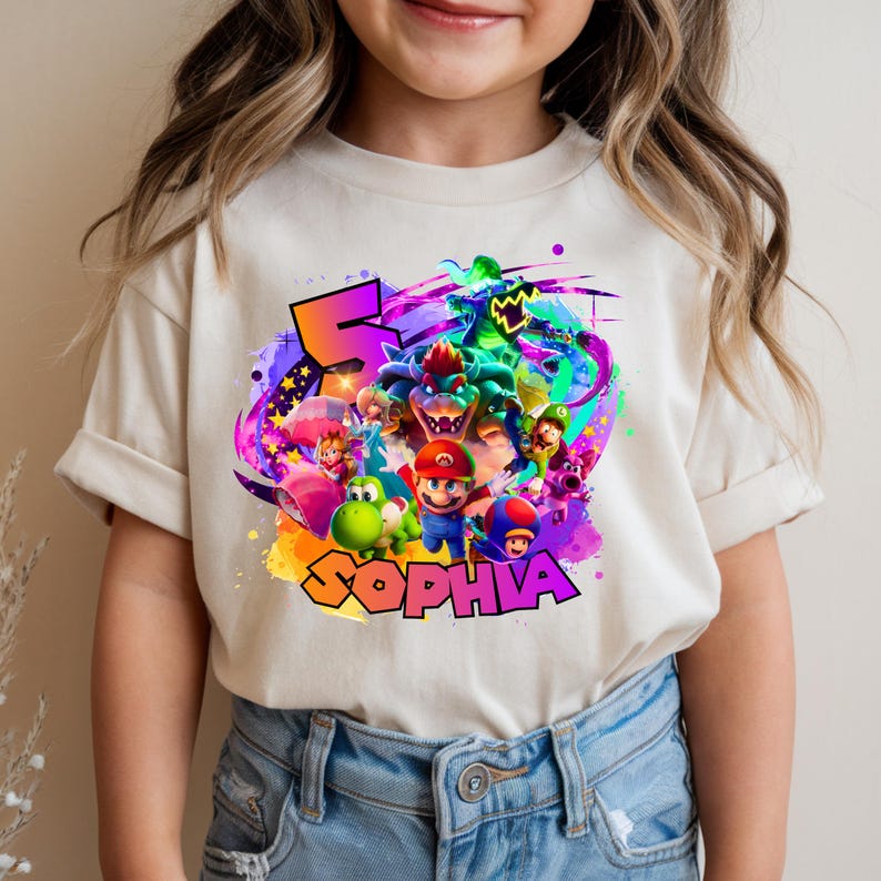 Puede incluir: Una camiseta de manga corta color crema con un estampado gr&aacute;fico colorido de Mario, Luigi, Yoshi, Bowser y Toad. El dise&ntilde;o incluye un n&uacute;mero 5 grande de color naranja y el nombre SOPHIA en letras moradas sobre un fondo de salpicaduras.