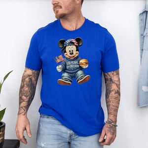May include: Royal blue t-shirt featuring a graphic of Mickey Mouse wearing a patriotic hat and sweater, holding a small American flag and a hamburger. The sweater reads "250 Years of Freedom".