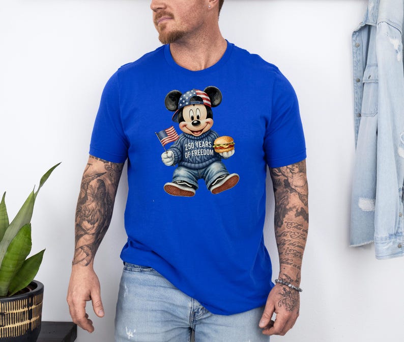 May include: Royal blue t-shirt featuring a graphic of Mickey Mouse wearing a patriotic hat and sweater, holding a small American flag and a hamburger. The sweater reads "250 Years of Freedom".