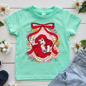 May include: A mint green t-shirt featuring a graphic of a red-haired princess within a floral and pearl-framed oval, accented with a red ribbon. The princess wears a purple top and is surrounded by colorful flowers. The shirt is paired with blue jeans.