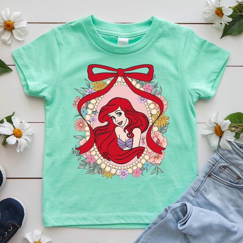 May include: A mint green t-shirt featuring a graphic of a red-haired princess within a floral and pearl-framed oval, accented with a red ribbon. The princess wears a purple top and is surrounded by colorful flowers. The shirt is paired with blue jeans.