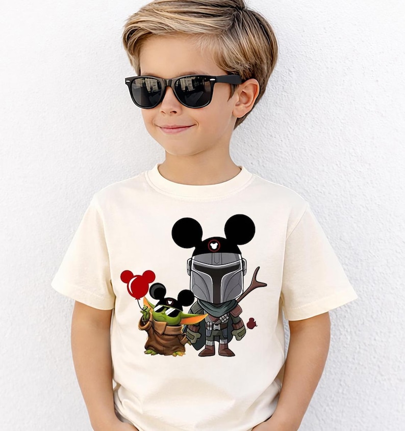 May include: Cream-colored t-shirt featuring a cartoon illustration of Mickey Mouse as The Mandalorian and Grogu. Mickey Mouse is wearing a Mandalorian helmet and Grogu is holding red balloons. The shirt is for children.