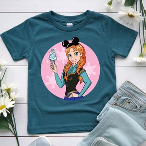 May include: Teal t-shirt featuring a cartoon illustration of a character with red hair, wearing a black top and blue skirt, holding a blue ice cream cone. The character is set against a pink circle with snowflake accents, and wears black mouse ears.