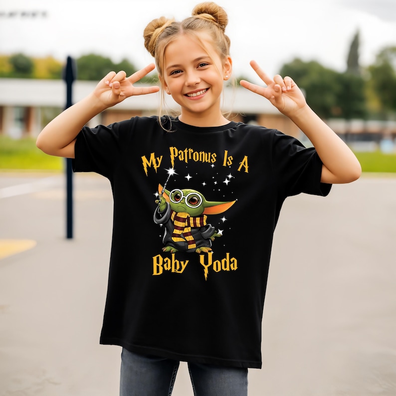May include: Black t-shirt with the text "My Patronus Is A Baby Yoda" in gold lettering. The graphic features a cartoon Baby Yoda character with glasses, a wand, and a scarf. The shirt is worn by a person.