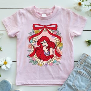 May include: A light pink t-shirt featuring a cartoon illustration of a mermaid with red hair, framed by a red ribbon and a floral border. The mermaid is wearing a purple top. The shirt is made of a soft material.