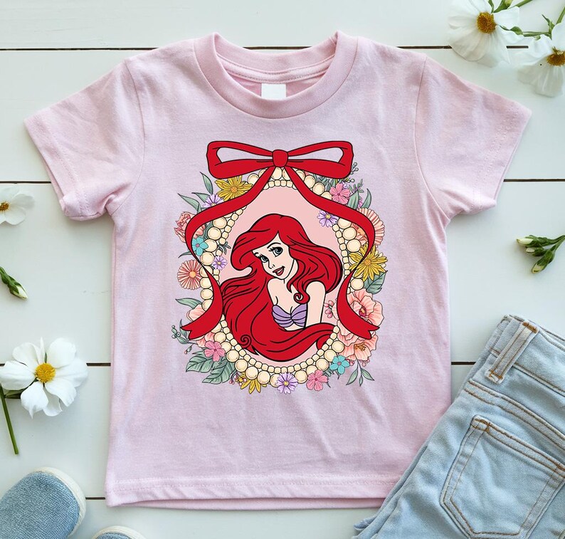 May include: A light pink t-shirt featuring a cartoon illustration of a mermaid with red hair, framed by a red ribbon and a floral border. The mermaid is wearing a purple top. The shirt is made of a soft material.