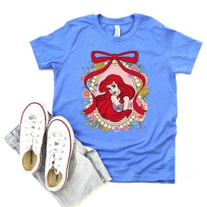 May include: A light blue t-shirt featuring a graphic of a red-haired mermaid within a floral and pearl frame, accented by a red bow. White sneakers and denim shorts are placed below the shirt.