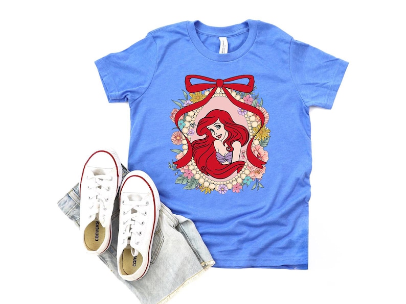 May include: A light blue t-shirt featuring a graphic of a red-haired mermaid within a floral and pearl frame, accented by a red bow. White sneakers and denim shorts are placed below the shirt.